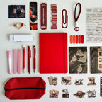 Collection of stationery items including a red notebook, pens, markers, and other office supplies on a white background.