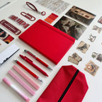 Red notebook, pens, markers, and photo album on a white surface with decorative items.