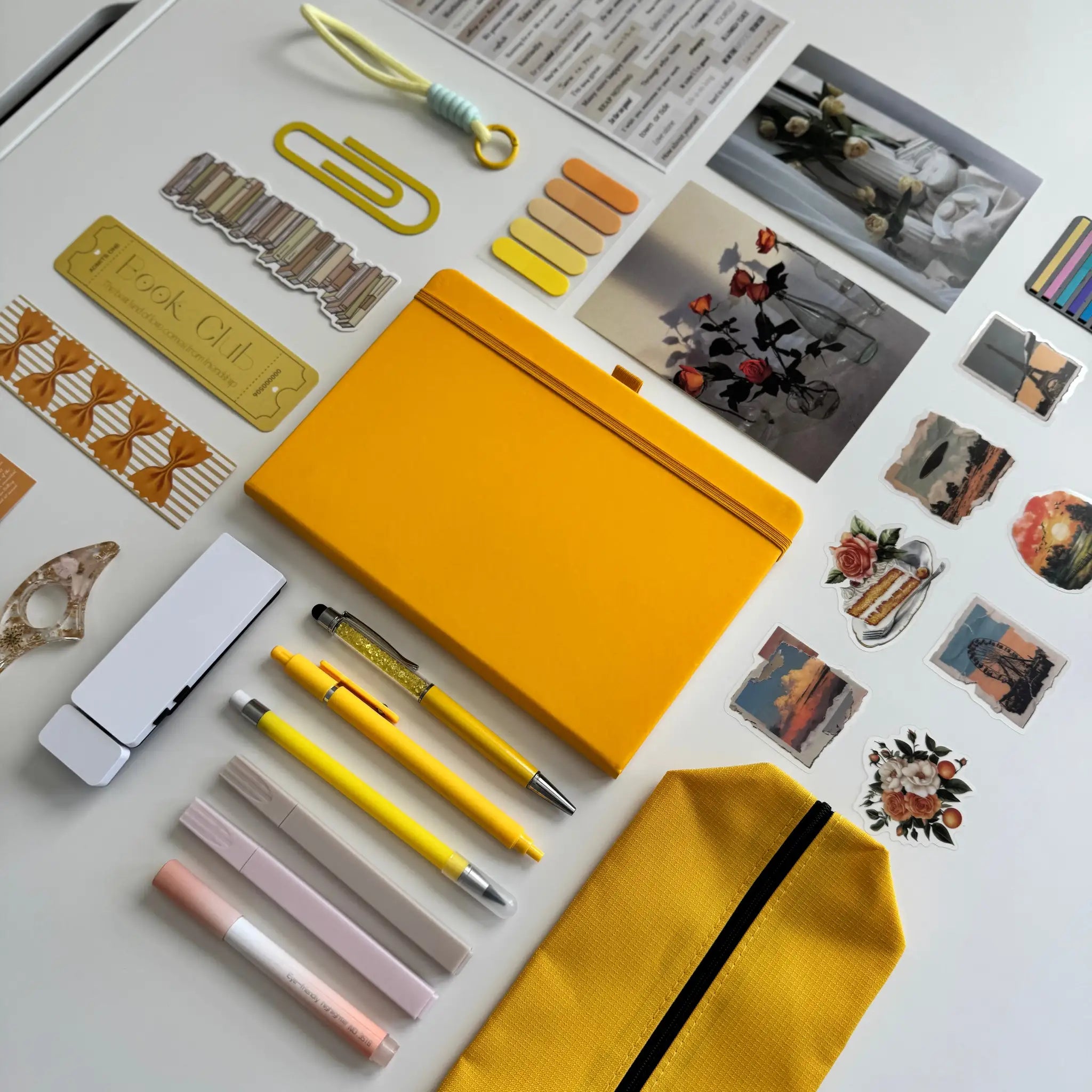 Collection of stationery items including a yellow notebook, pens, and decorative elements on a light surface.