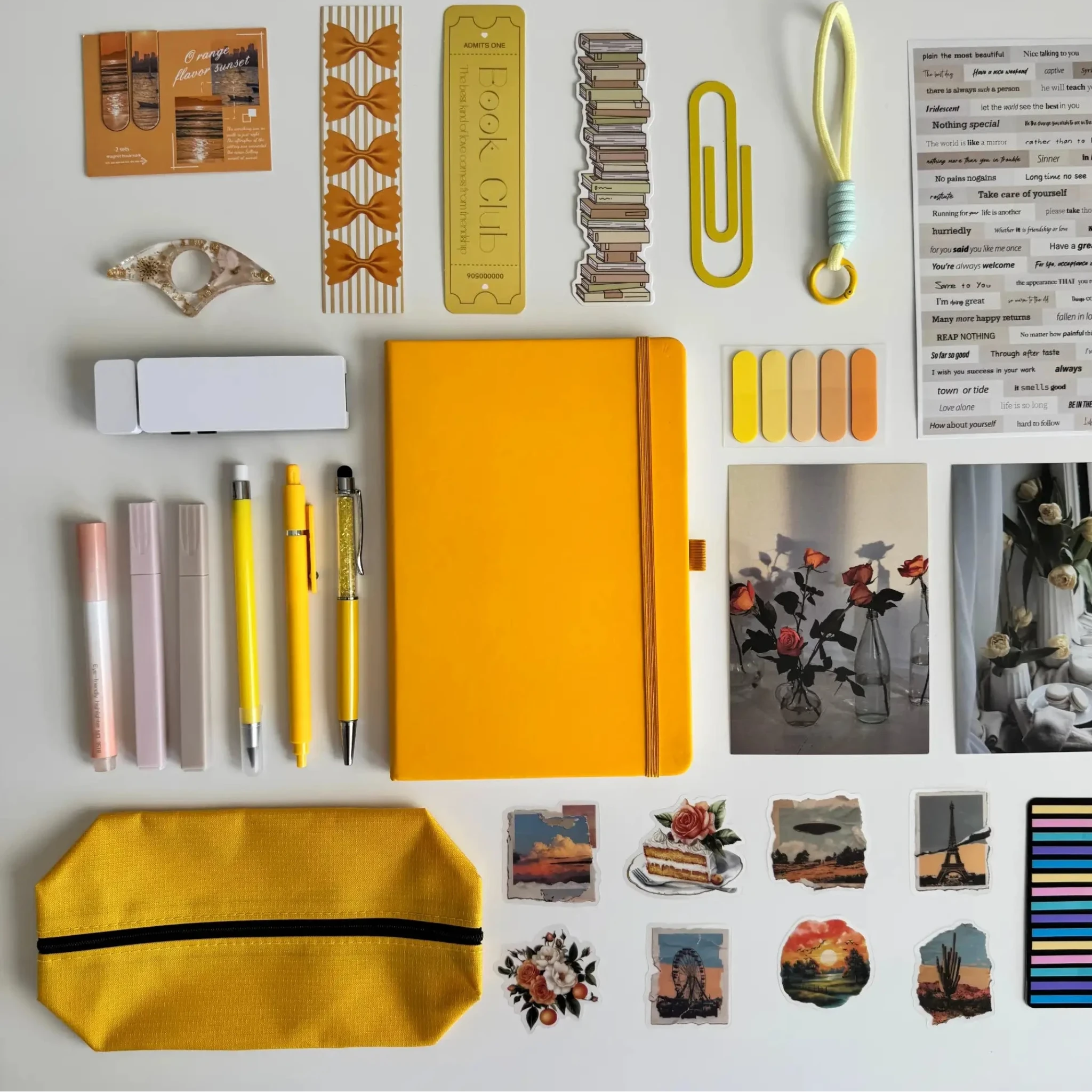 Collection of stationery items including a yellow notebook, pens, markers, and decorative elements on a white background.
