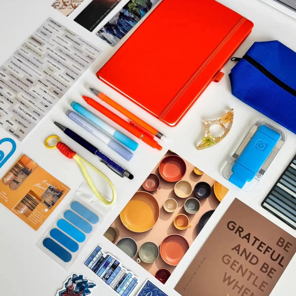 Collection of stationery items including a  notebook, pens, markers, and photo prints in blue and orange colors on a light background