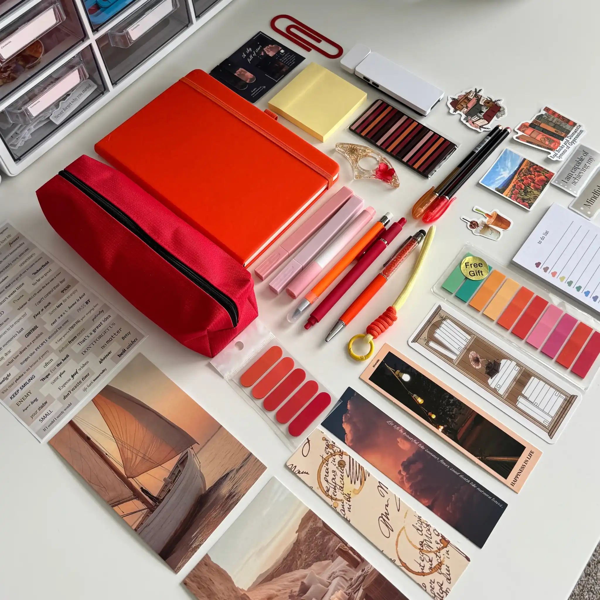 A styled flat-lay of stationery and accessories arranged on a white surface, including a red zip pouch, orange notebook, pens, sticky notes, bookmarks, cards, and small decorative items.