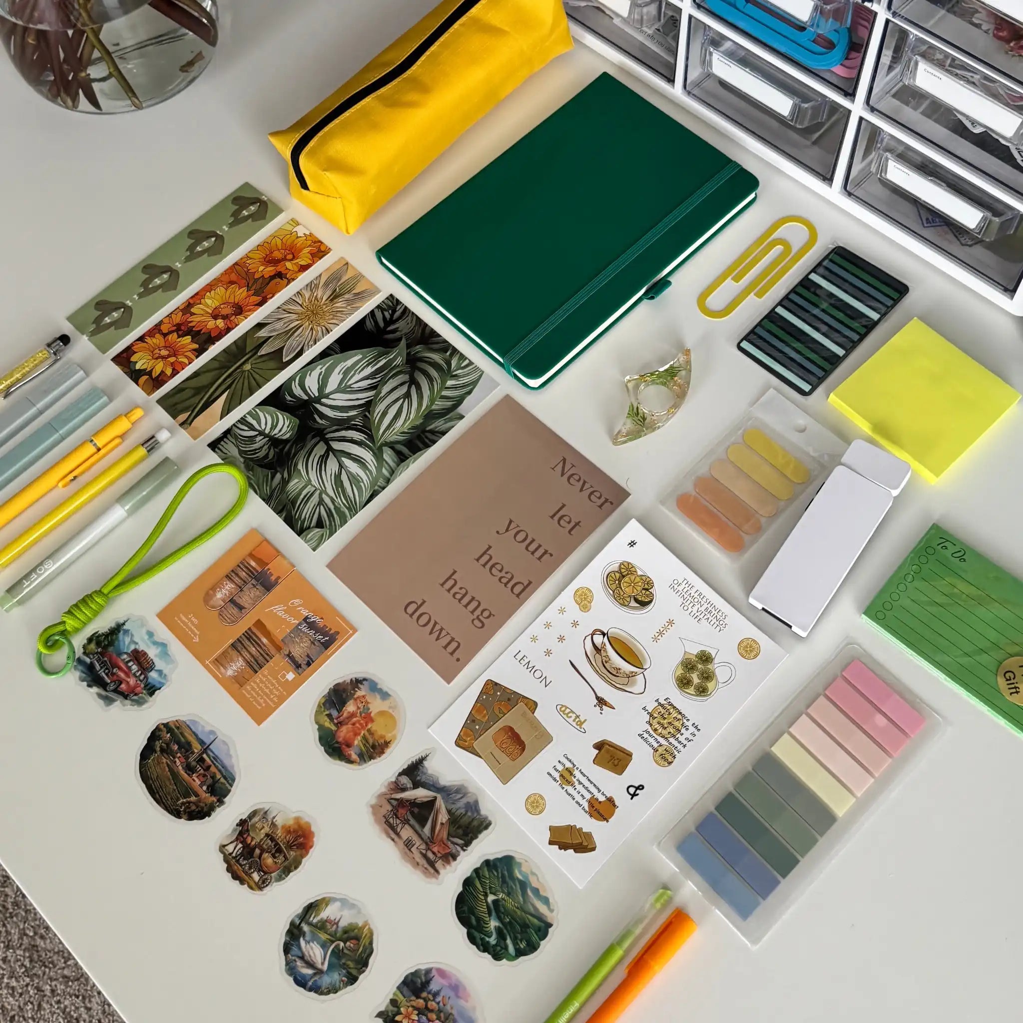 A styled flat-lay of a stationery and desk accessories set arranged on a white tabletop, with a green notebook, yellow pencil case, stickers, pens, sticky notes, and small accessories. A vase of pink roses and a white organizer filled with supplies are visible in the background.