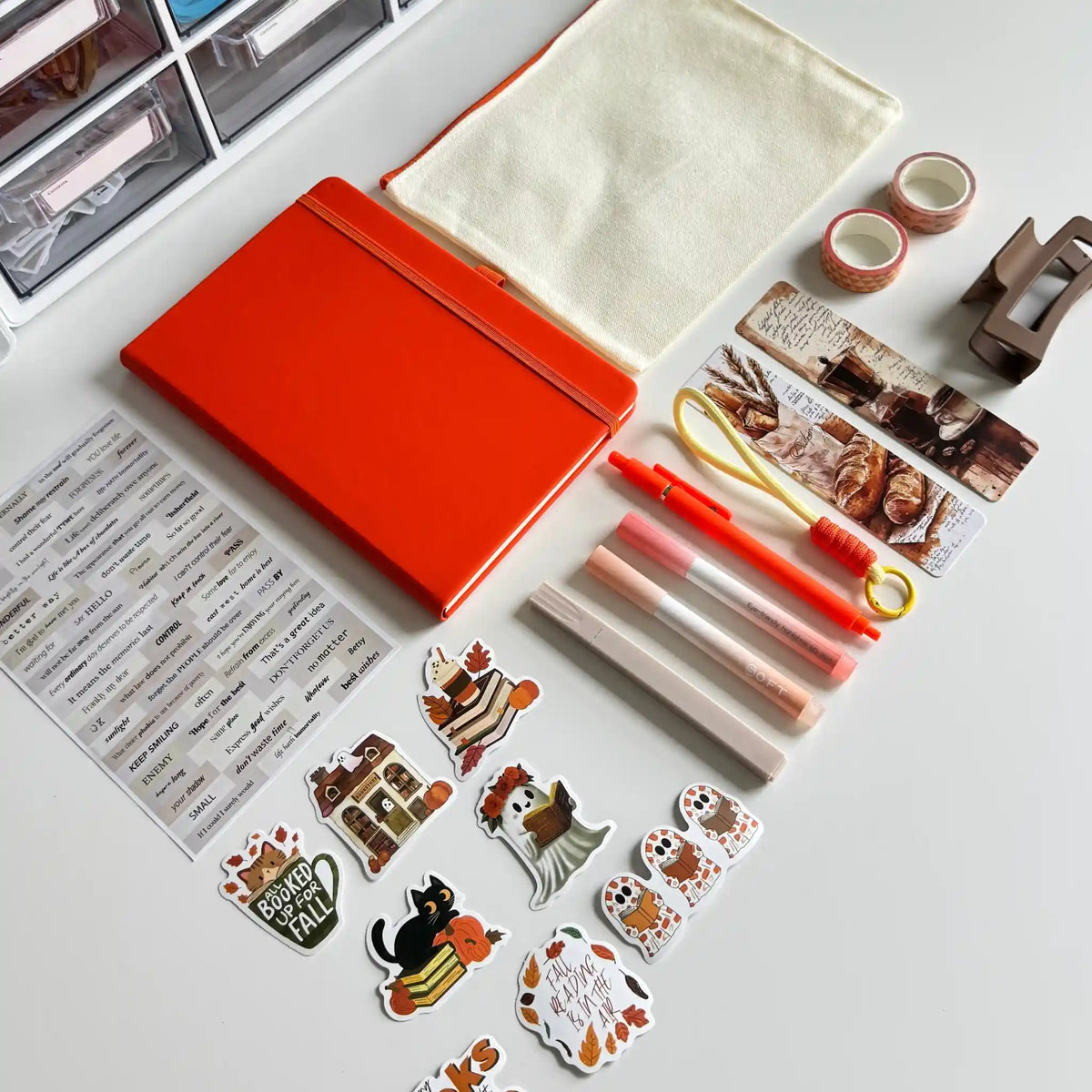 A styled flat-lay of a stationery and desk accessory kit on a white tabletop, including a bright orange notebook, a cream fabric pouch, sticker sheets with animal illustrations, pens, washi tape, tape dispenser, and organizer drawers in the background.