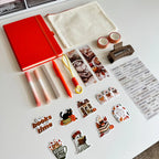 A styled flat-lay of stationery and desk accessories on a white surface, including a bright red notebook, a cream fabric pouch, washi tapes, highlighters, bookmarks, a small brown hair clip, a sticker sheet, and multiple autumn-themed stickers.
