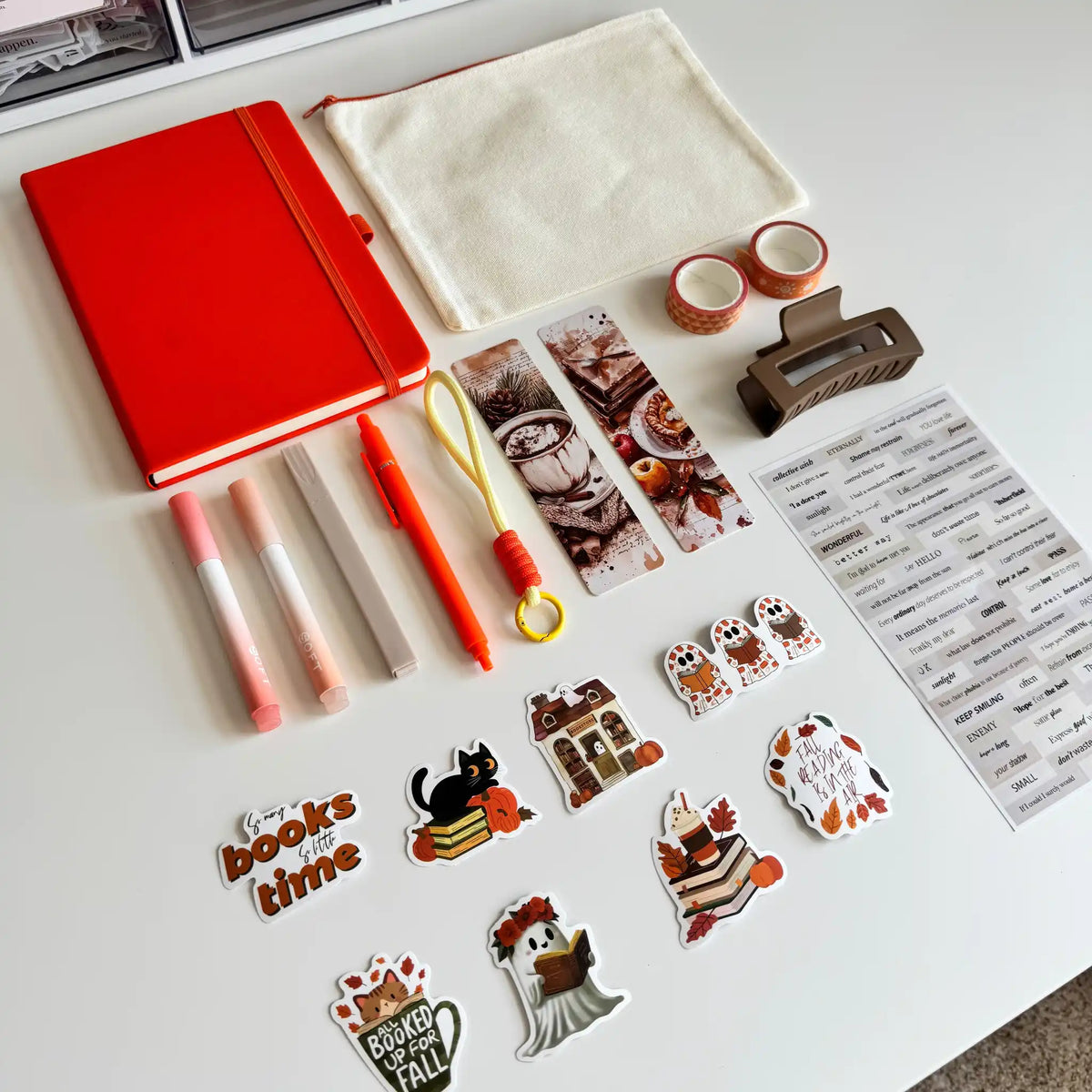 A styled flat-lay of stationery and desk accessories on a white surface, including a bright red notebook, a cream fabric pouch, washi tapes, highlighters, bookmarks, a small brown hair clip, a sticker sheet, and multiple autumn-themed stickers.