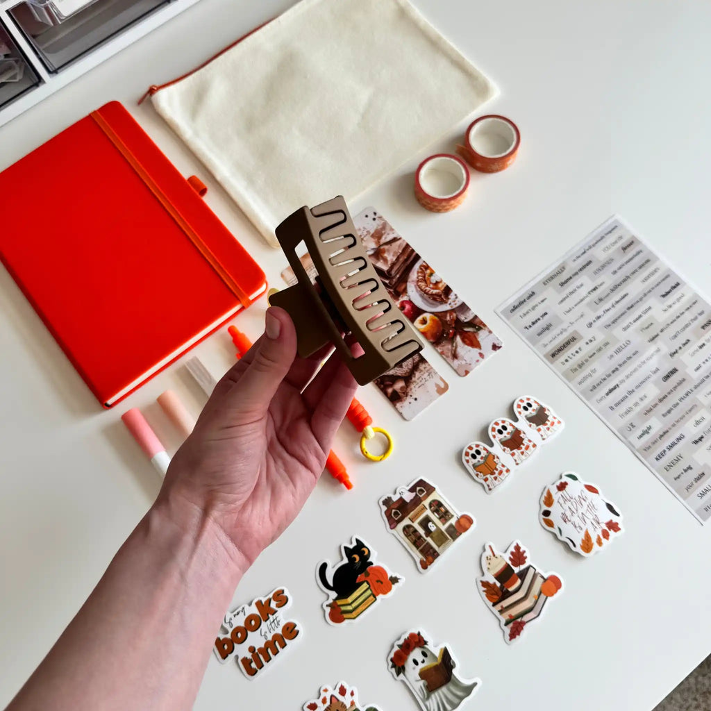 A hand holds a brown claw clip decorated with autumn-themed stickers, surrounded by matching stationery items on a white desk, including a red notebook, a cream pouch, washi tape, and sticker sheets.
