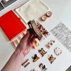 A hand holds a brown claw clip decorated with autumn-themed stickers, surrounded by matching stationery items on a white desk, including a red notebook, a cream pouch, washi tape, and sticker sheets.