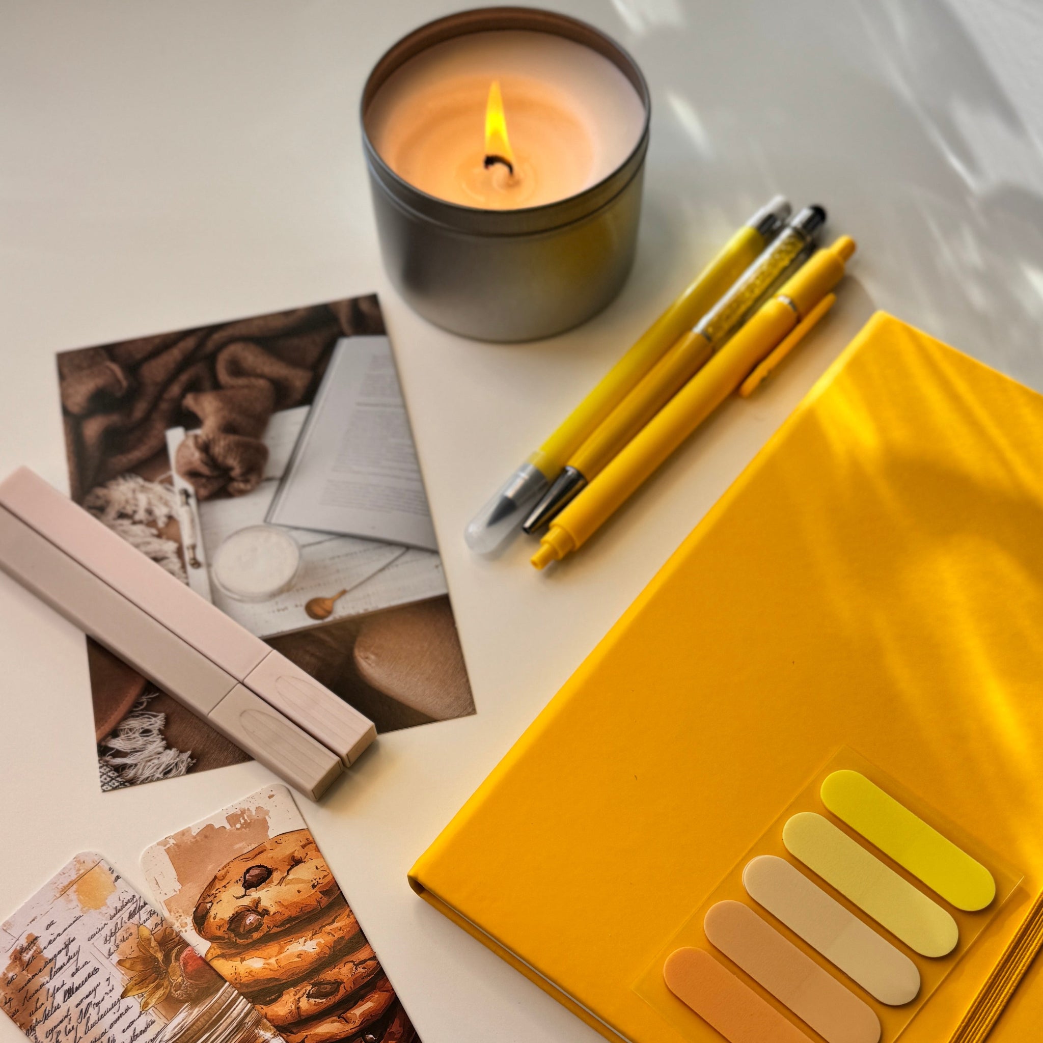 Yellow notebook with color swatches, two pens, a lit candle, and a magazine on a white surface.