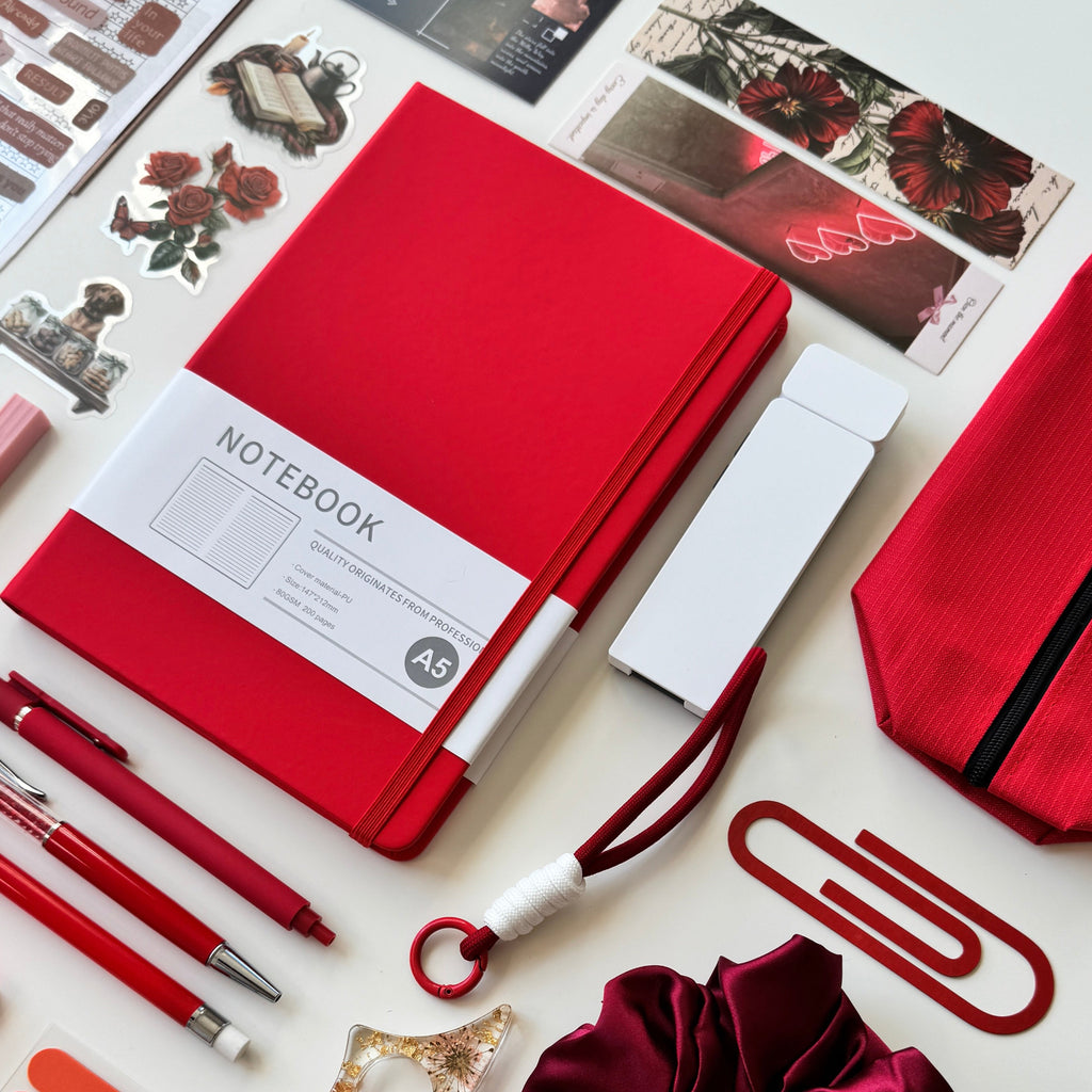 Collection of stationery items including a red notebook, pen, scrunchie, stickers and bookmarks on a white background.