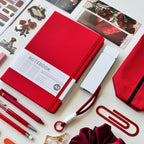 Collection of stationery items including a red notebook, pen, scrunchie, stickers and bookmarks on a white background.