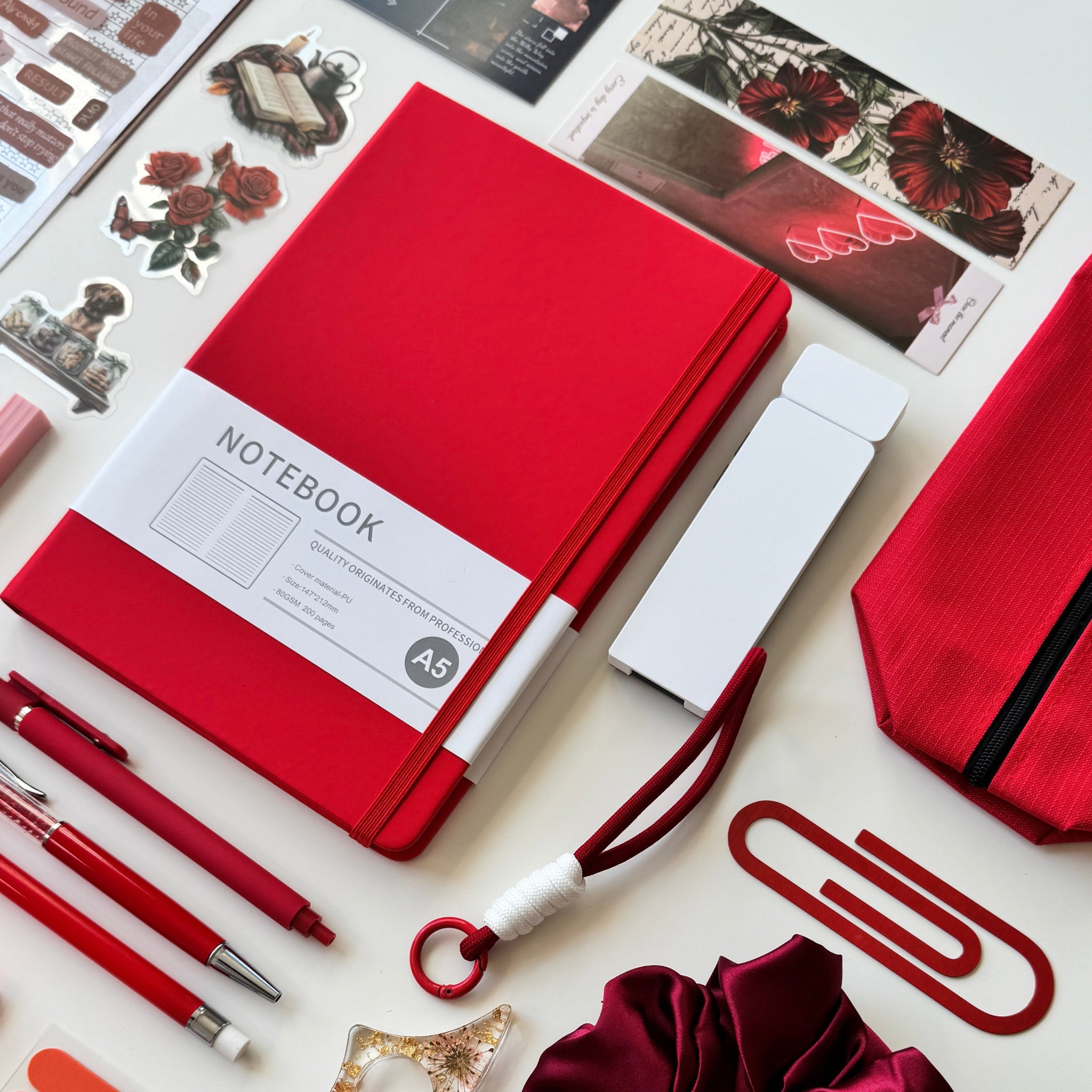Collection of stationery items including a red notebook, pen, scrunchie, stickers and bookmarks on a white background.
