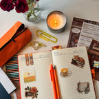 Notebook with orange pen, candle, and flowers on a desk
