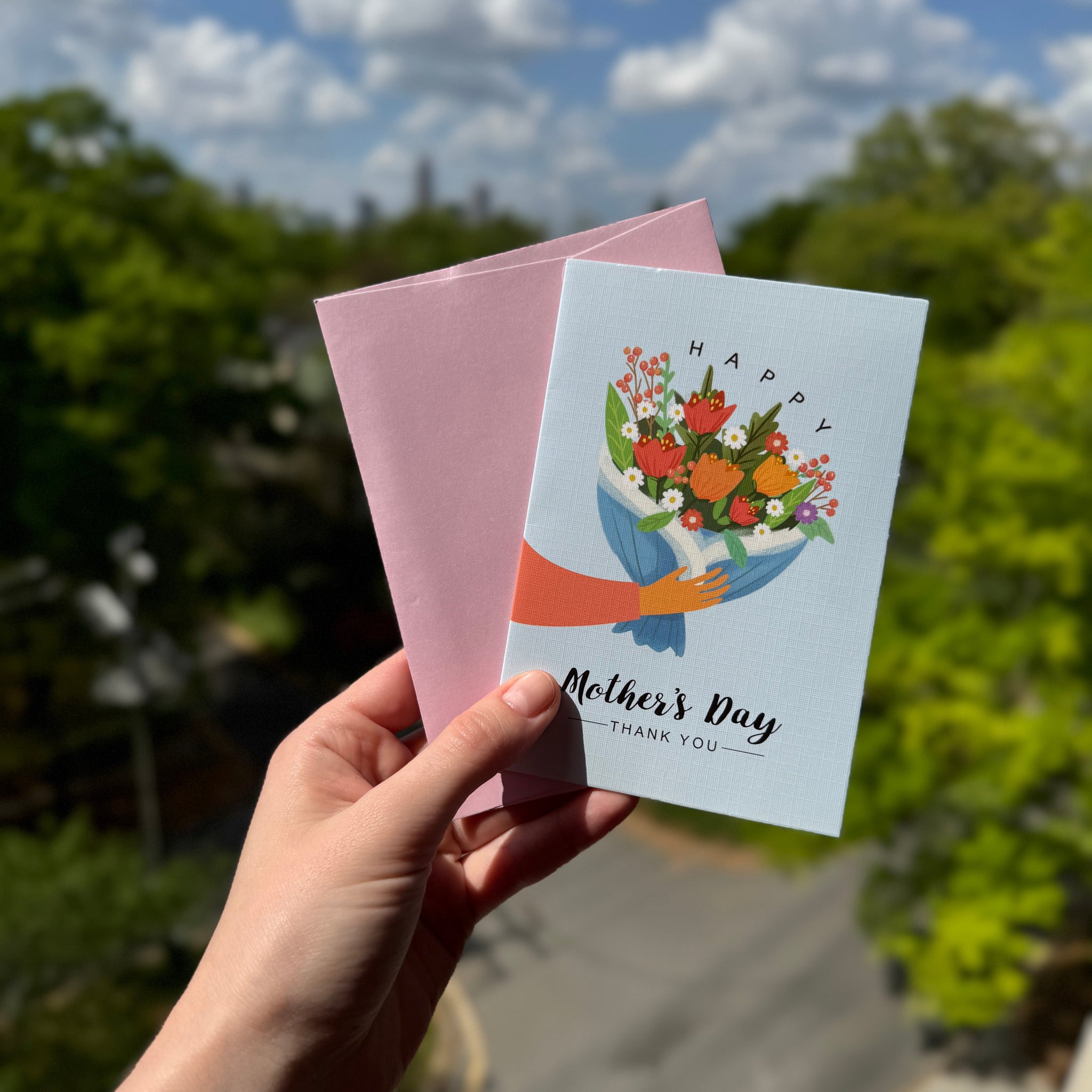 Hand holding a 'Happy Mother's Day' card with a floral design outdoors.
