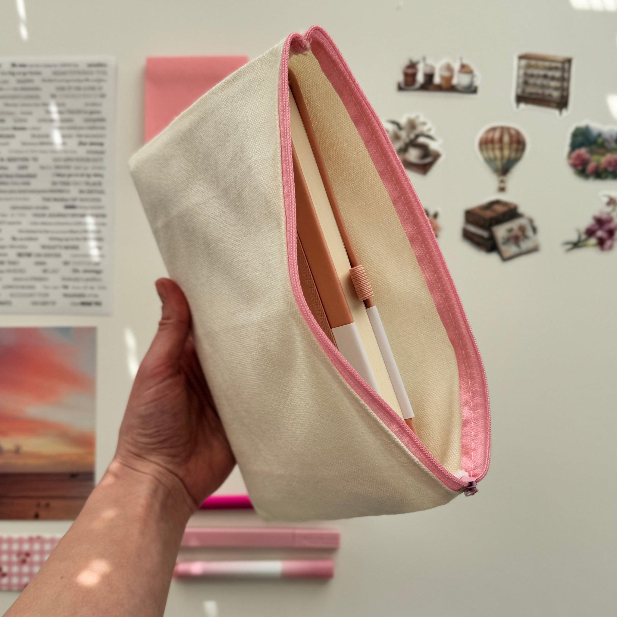 Hand holding a beige clutch with pink trim against a wall with photos and notes.