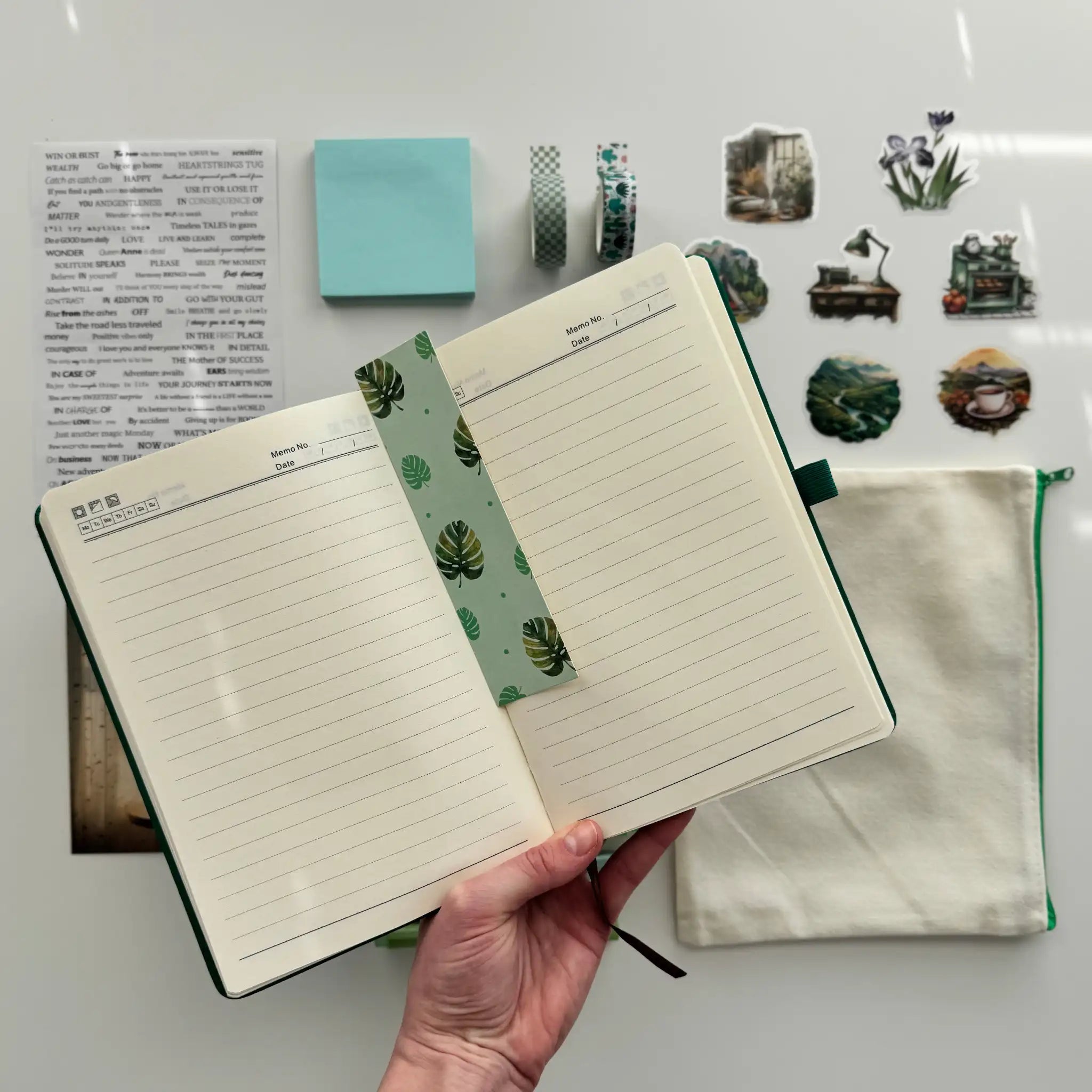 Open notebook with lined pages held by a hand, surrounded by stationery items on a light gray background.
