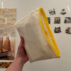 Hand holding a beige pouch with a yellow edge in front of a refrigerator with magnets and photos.