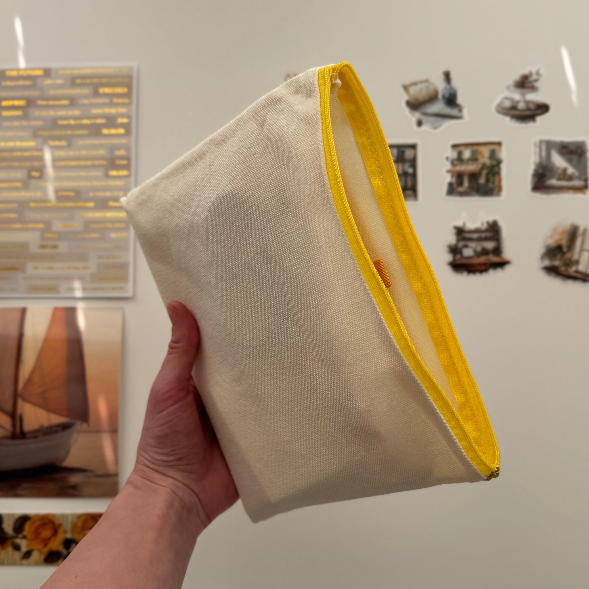 Hand holding a beige pouch with a yellow edge in front of a refrigerator with magnets and photos.