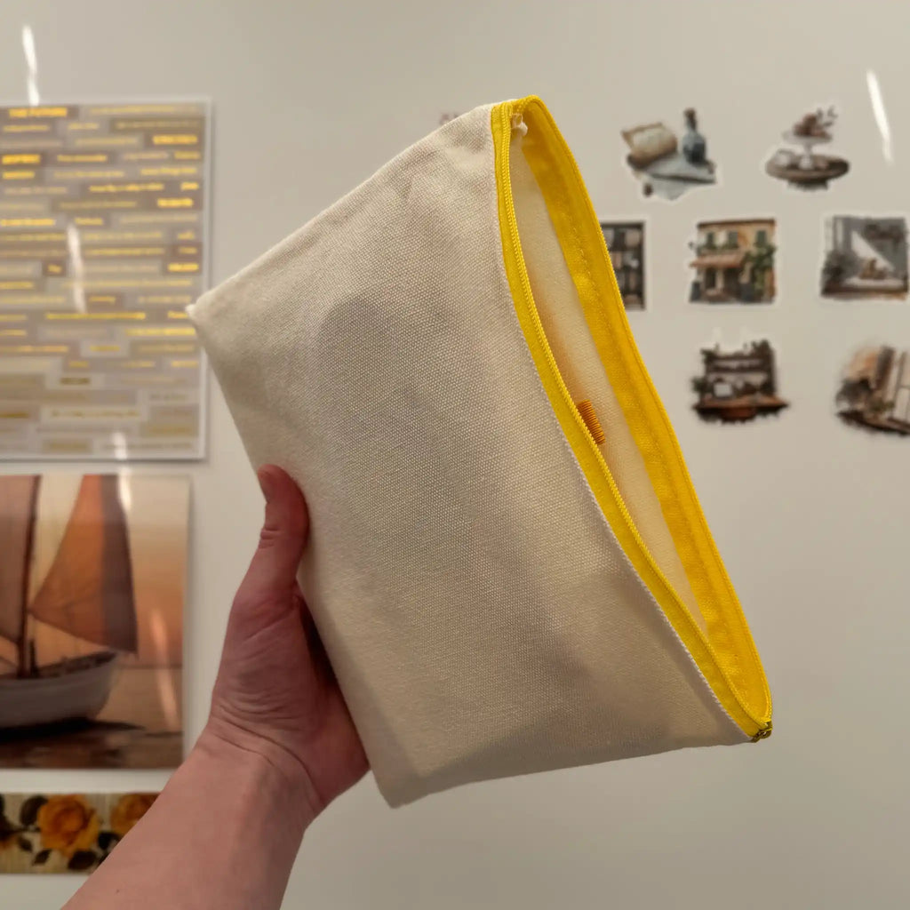 Hand holding a beige pouch with yellow edge against a wall with framed pictures