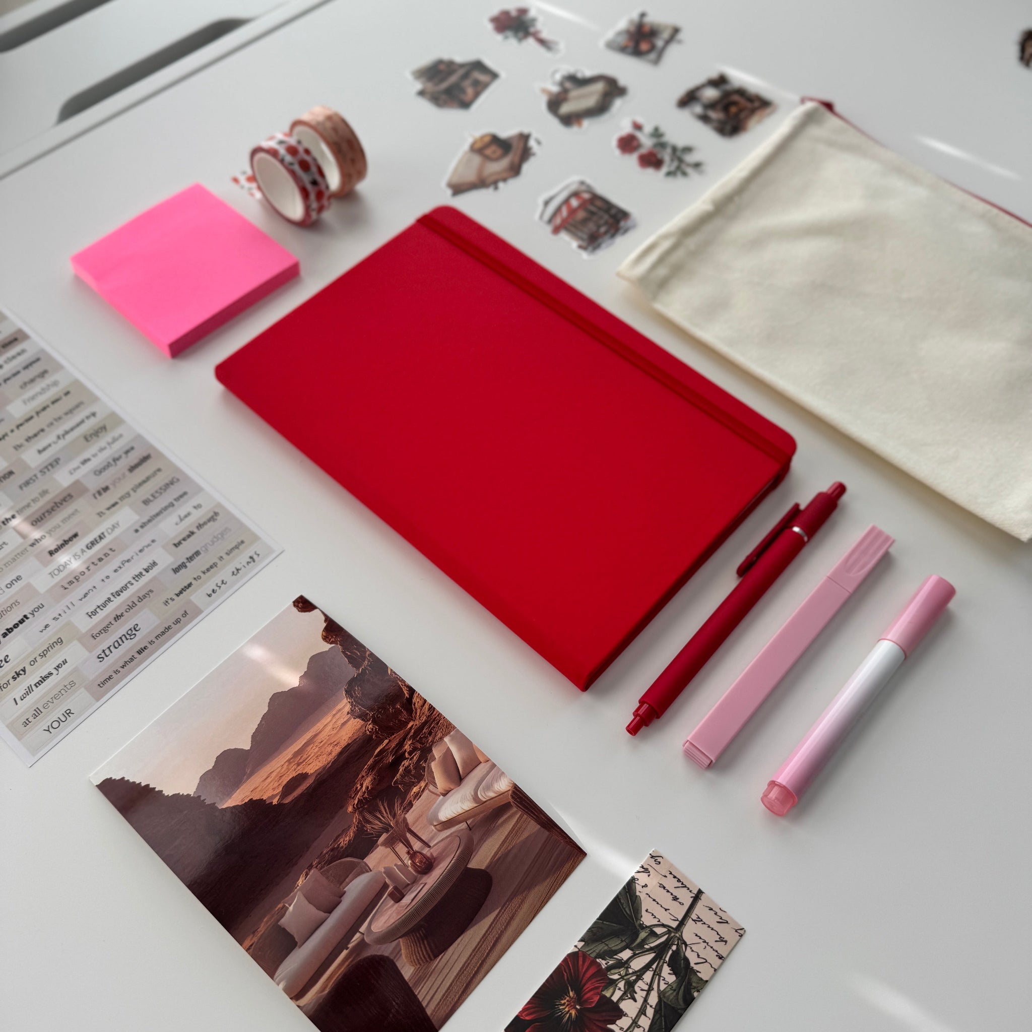 Red notebook, pink and red pens, and other stationery items on a white surface.