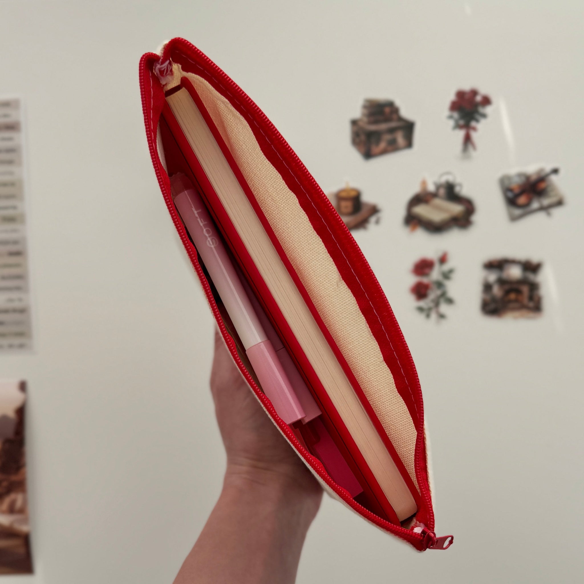 Hand holding a red pouch with a book inside against a white wall with decorative elements.