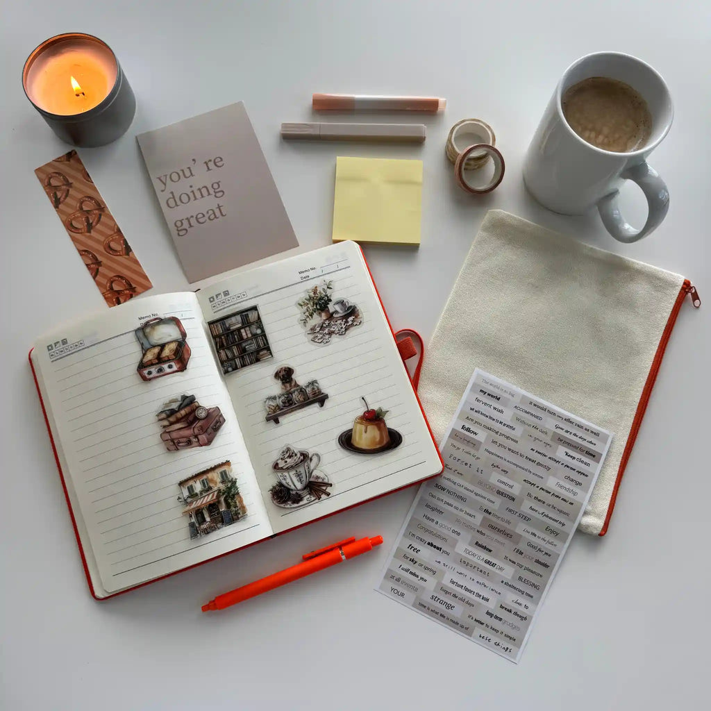 Open notebook with stickers, a mug of coffee, a candle, and stationery items on a white surface.