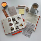 Open notebook with stickers, a mug of coffee, a candle, and stationery items on a white surface.