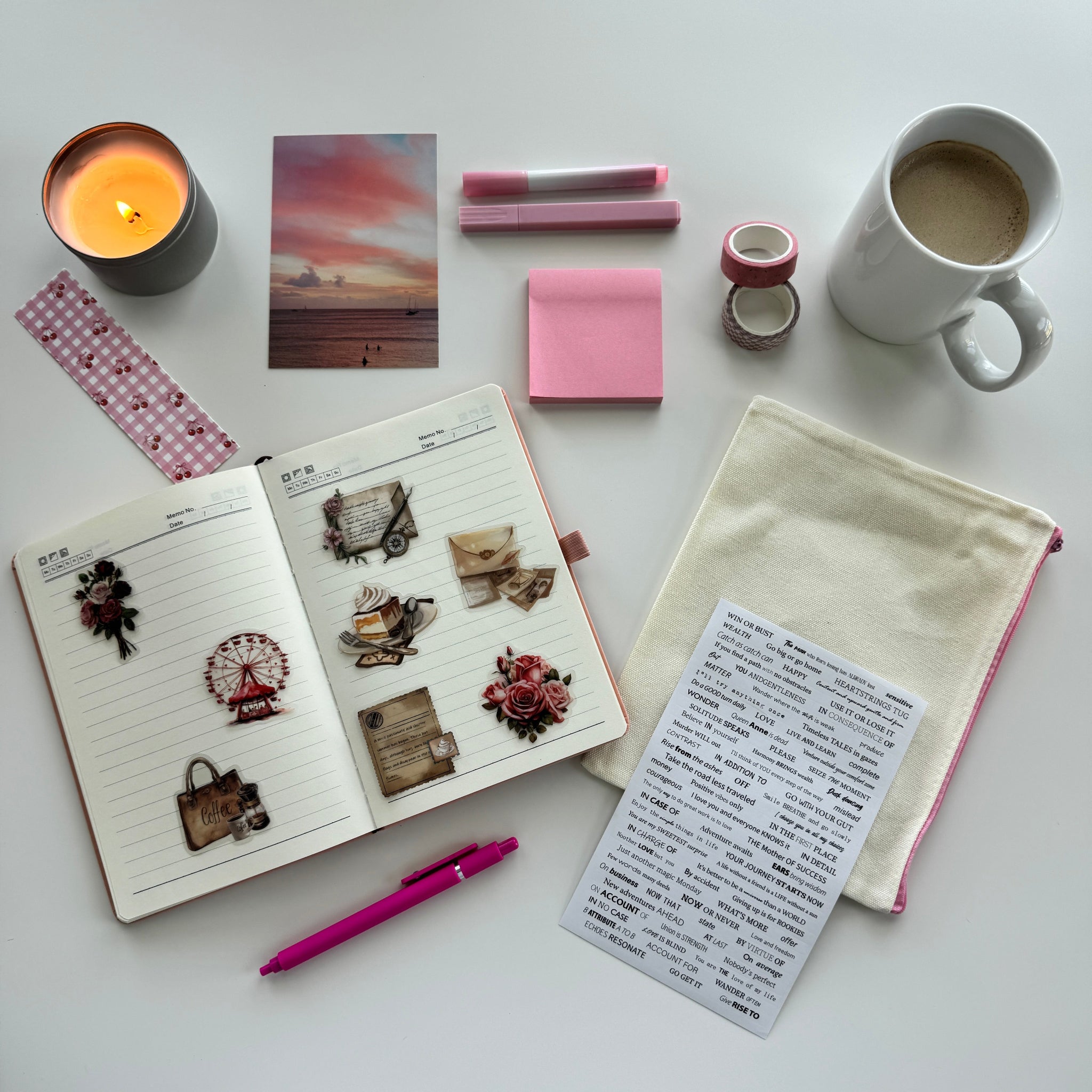 Desk with open notebook, pink pen, candle, mug, and stationery items on a white surface
